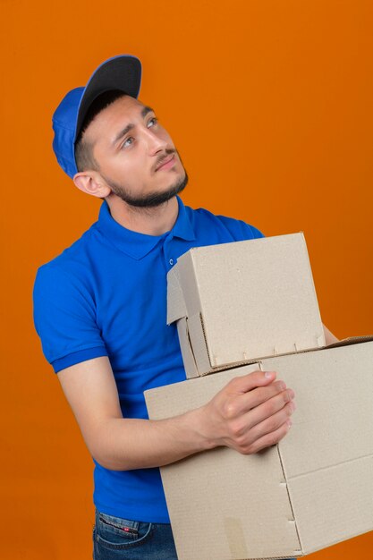 Junger nachdenklicher Lieferbote, der blaues Poloshirt und Mütze trägt, die mit Stapel von Kisten stehen, sehen verträumt über lokalisierten orange Hintergrund