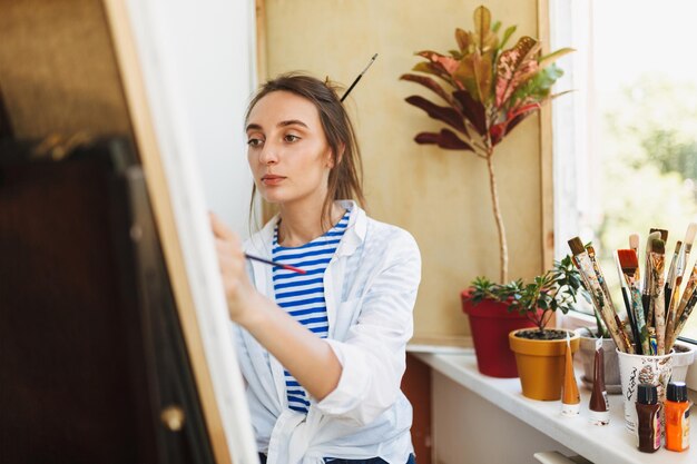 Junger nachdenklicher Maler in weißem Hemd und gestreiftem T-Shirt, der zu Hause verträumt auf Staffelei in der Nähe des Fensters zeichnet