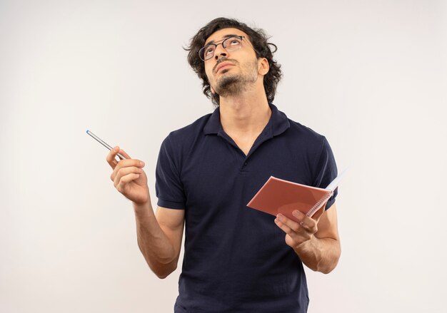 Junger nachdenklicher Mann im schwarzen Hemd mit optischer Brille hält Stift und Notizbuch lokalisiert auf weißer Wand