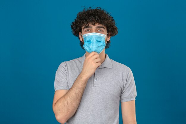 Junger nachdenklicher Mann in der medizinischen Schutzmaske mit Hand auf Kinn nachdenklichem Blick, der über lokalisiertem blauem Hintergrund steht