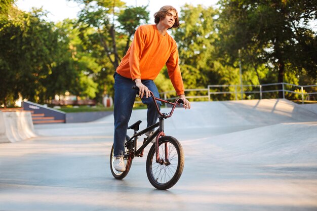 Junger nachdenklicher Typ in orangefarbenem Pullover und Jeans, der verträumt Fahrrad im modernen Skatepark fährt