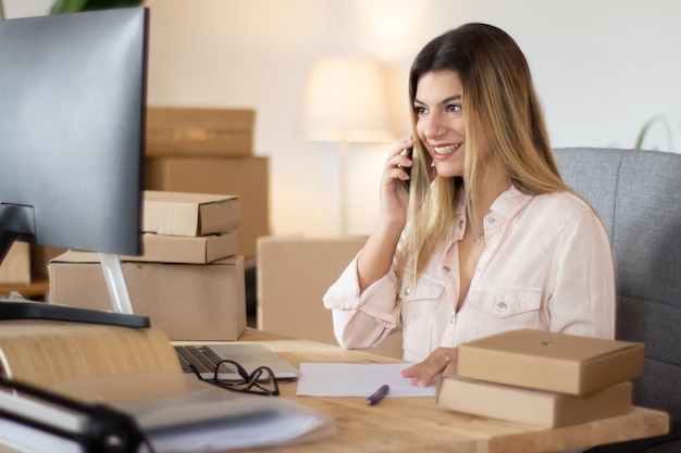 Junger Online-Verkäufer berät Kunden am Telefon, schaut auf Computerbildschirm und lächelt. Schöne kaukasische Frau, die im Heimbüro arbeitet, umgeben von Papppaketen. E-Commerce-Konzept