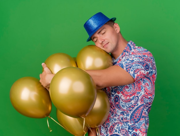 Kostenloses Foto junger party-typ mit geschlossenen augen, der blauen hut trägt, der neben luftballons steht, die auf grün lokalisiert werden