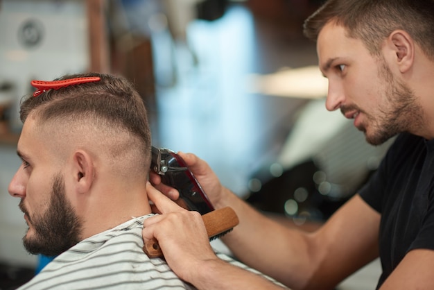 Junger professioneller männlicher Friseur, der sich konzentriert, während er seinem Kunden einen Haarschnitt gibt.