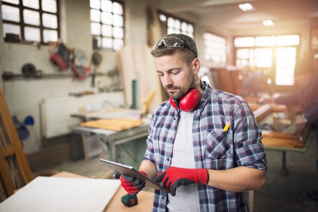 Junger professioneller Tischler mit Schutzbrille, der Tablet-Computer hält und Entwurf seines Projekts in der Werkstatt prüft