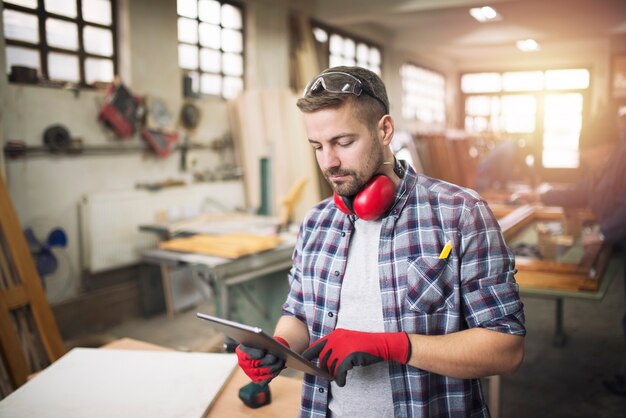 Junger professioneller Tischler mit Schutzbrille, der Tablet-Computer hält und Entwurf seines Projekts in der Werkstatt prüft