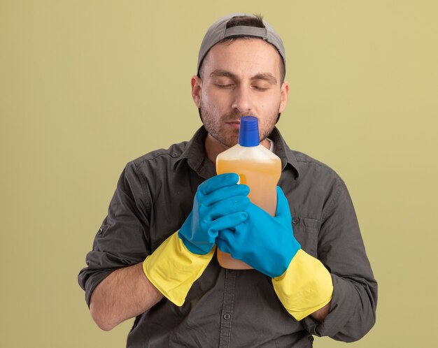 Junger Reinigungsmann, der Freizeitkleidung und Kappe in den Gummihandschuhen hält, die Flasche mit Reinigungsmitteln halten, die frisches Aroma einatmen, das über grüner Wand steht