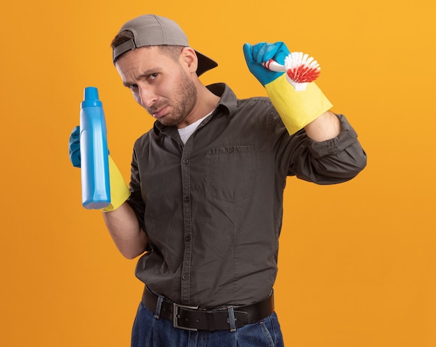 Junger Reinigungsmann, der Freizeitkleidung und Kappe in den Gummihandschuhen trägt, die Reinigungsbürste und -flasche mit Reinigungsmitteln halten, die verwirrt und unzufrieden stehen über orange Wand stehen