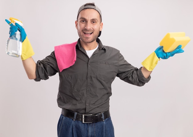 Junger Reinigungsmann, der Freizeitkleidung und Mütze in Gummihandschuhen trägt, die Reinigungsspray und Schwamm mit Lappen auf seiner Schulter halten, die glücklich und aufgeregt stehen, über orange Wand stehend