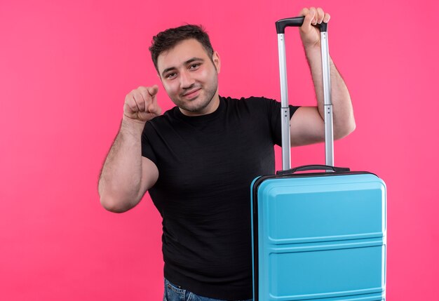 Junger Reisender im schwarzen T-Shirt, das Koffer glücklich und positiv zeigt mit Zeigefinger hält, lächelt und über rosa Wand steht