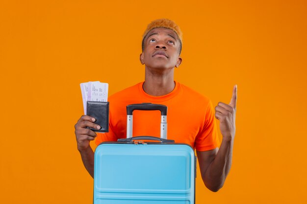 Junger reisender Junge, der orange T-Shirt hält, das Flugtickets und Koffer schaut, die nach oben schauen und auf etwas mit ernstem Gesicht zeigen, das über orange Wand steht