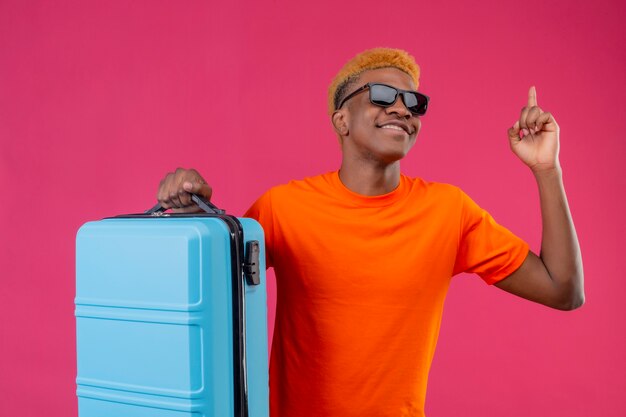 Junger reisender Junge, der orange T-Shirt hält Koffer hält, der mit dem Finger oben zeigt und lächelnd über rosa Wand stehend schaut