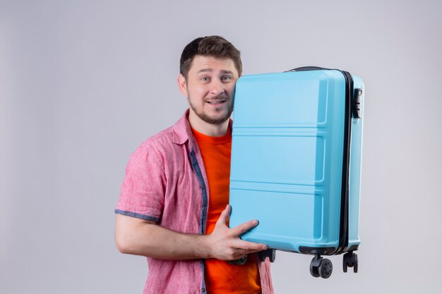 Junger reisender Mann, der blauen Koffer hält, der Kamera betrachtet, die fröhlich positiv und glücklich steht, die über weißem Hintergrund 2 steht