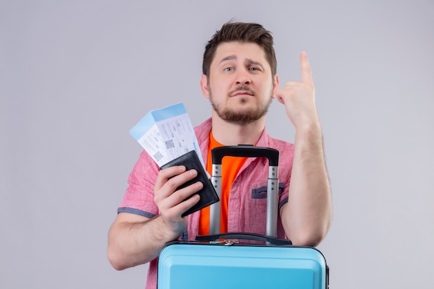 Junger reisender Mann, der Flugtickets und Koffer hält, die zuversichtlich zeigen, mit dem Finger oben über lokalisiertem weißem Hintergrund stehend