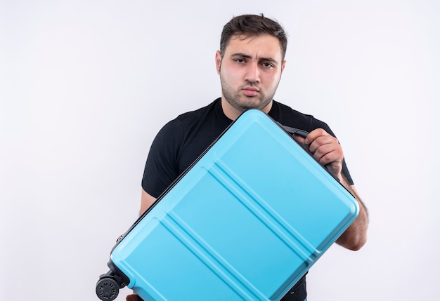 Junger reisender Mann im schwarzen T-Shirt, das Koffer mit ernstem Stirnrunzeln hält, das über weißer Wand steht