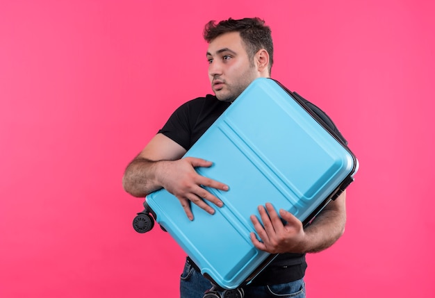 Kostenloses Foto junger reisender mann im schwarzen t-shirt, der koffer hält, der beiseite schaut, verwechselt mit angstausdruck, der über rosa wand steht