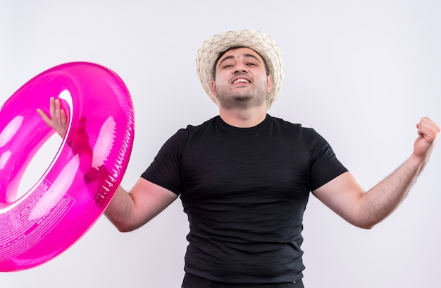 Junger reisender Mann im schwarzen T-Shirt und im Sommerhut, der aufblasbare Ringballenfäuste hält, die sich über seinen Erfolg freuen, der über weißer Wand steht