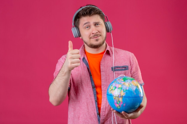 Junger reisender Mann mit Kopfhörern, die Musik hören, die Globus zeigt, der Daumen oben lächelnd über rosa Hintergrund steht