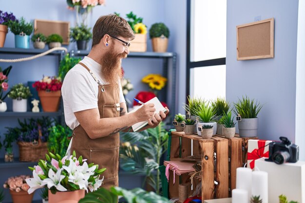 Junger rothaariger Mann, Florist, lächelt selbstbewusst und schreibt auf Notizbuch im Blumenladen