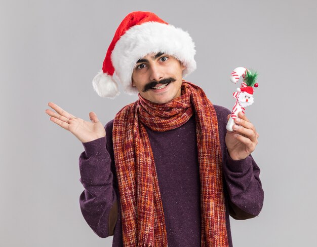 Junger schnurrbärtiger Mann mit Weihnachtsmütze mit warmem Schal um den Hals, der Weihnachtszuckerstange glücklich und fröhlich lächelnd über weißer Wand hält