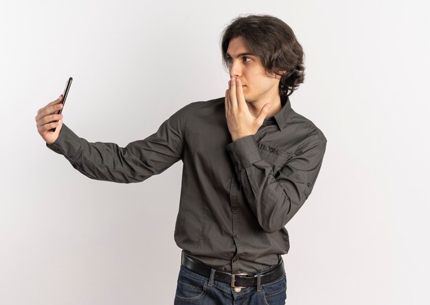 Junger schockierter hübscher kaukasischer Mann legt Hand auf Mund und betrachten Telefon lokalisiert auf weißem Hintergrund mit Kopienraum