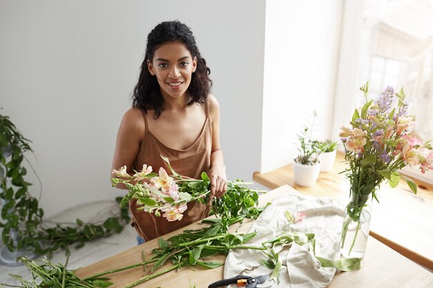 Junger schöner afrikanischer Frauenflorist, der Blumen am Arbeitsplatz über weißer Wand kümmert.