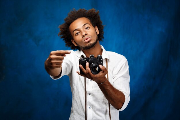 Junger schöner afrikanischer Mann, der alte Kamera über blauer Wand hält.