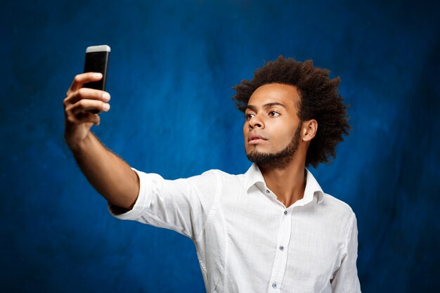 Junger schöner afrikanischer Mann, der selfie über blaue Wand macht.