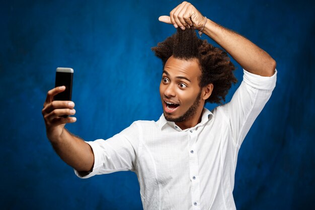 Junger schöner afrikanischer Mann, der selfie über blaue Wand macht.