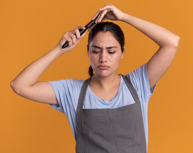 Junger schöner Frauenfriseur in der Schürze, die Haarbürste hält, die ihr Haar kämmt, das über orange Wand steht