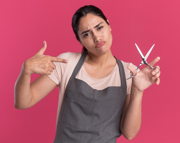 Junger schöner Frauenfriseur in der Schürze, die Schere hält, die mit Zeigefinger darauf zeigt und zuversichtlich steht über rosa Wand