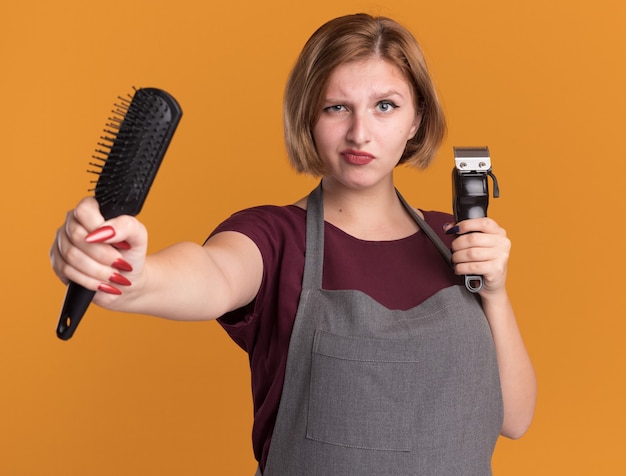 Junger schöner Frauenfriseur in der Schürze, die Trimmer und Haarbürste hält, die Front betrachtet, das schiefes Mund macht, das missfällt, das über orange Wand steht