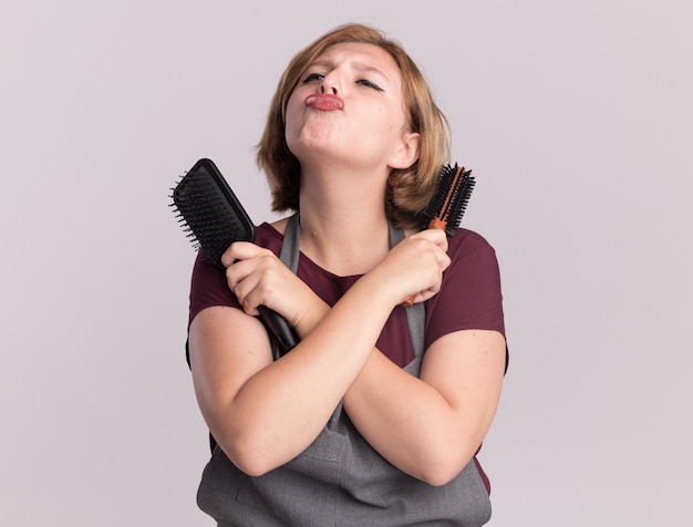 Junger schöner Fraufriseur in der Schürze, die Haarbürsten hält, die Hände kreuzen, die blasenden Kuss schauen, der über weißer Wand steht