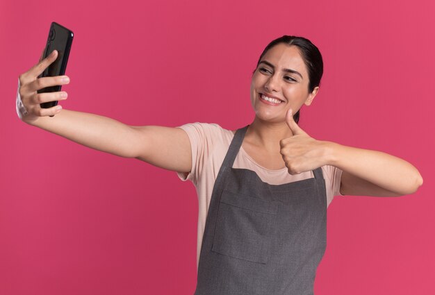 Junger schöner Fraufriseur in der Schürze, die Smartphone hält, das es tut, das selfie tut, das Daumen oben zeigt lächelnd steht über rosa Wand