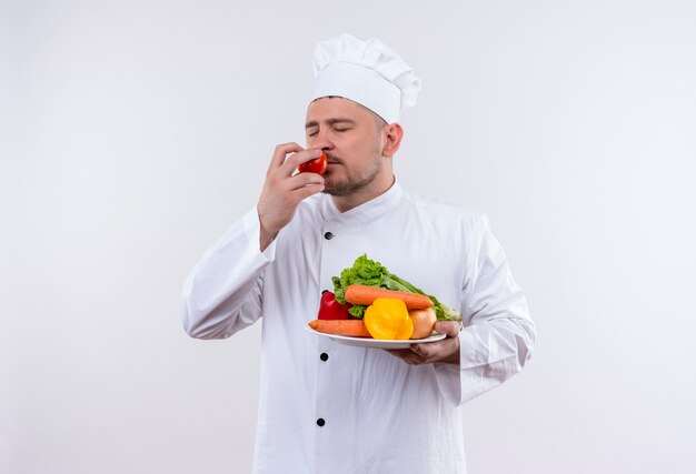 Junger schöner Koch in der Kochuniform, die Platte mit Gemüse und schnüffelnde Tomate mit geschlossenen Augen auf lokalisiertem weißen Raum hält