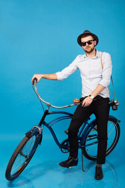 Junger schöner lächelnder glücklicher Mann, der auf Hipster-Fahrrad reist, das auf blauem Studiohintergrund aufwirft, Hemd, Hut und Sonnenbrille tragend