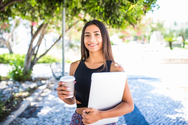 Junger schöner lateinamerikanischer Student mit Laptop und Tasse Kaffee, die im Park studieren. Mädchen geht im Park mit großem Lächeln und hält Laptop.