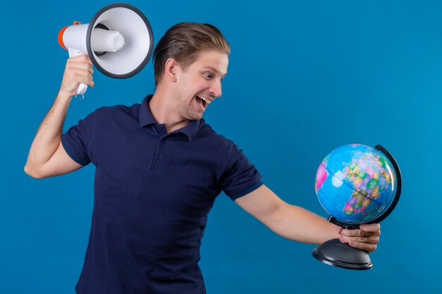 Junger schöner Mann, der Globus und Megaphon hält, die spielerisch und glücklich stehen über blauem Hintergrund stehen