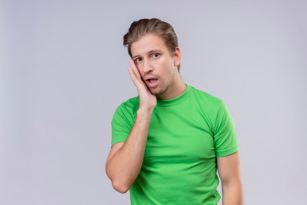 Junger schöner Mann, der grünes T-Shirt trägt, das mit Hand nahe Gesicht schaut