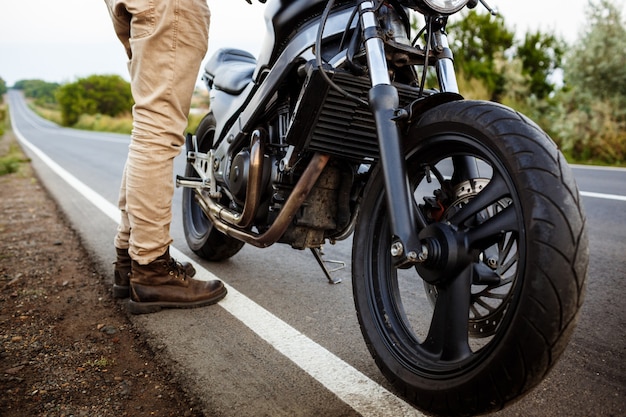 Junger schöner Mann, der nahe seinem Motorrad an der Landstraße aufwirft.