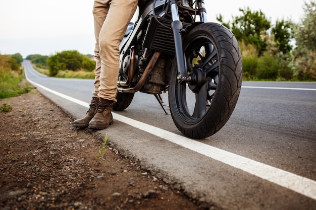 Junger schöner Mann, der nahe seinem Motorrad an der Landstraße aufwirft.