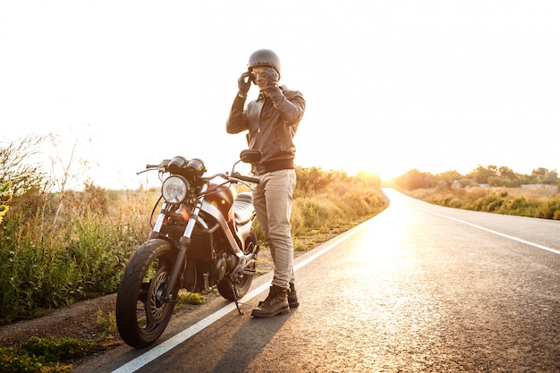 Junger schöner Mann, der nahe seinem Motorrad an der Landstraße aufwirft.