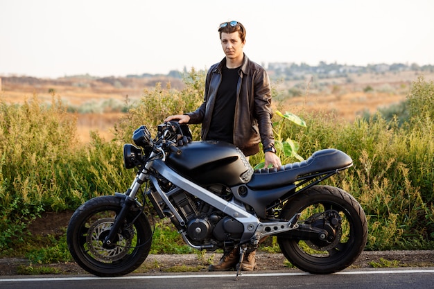 Kostenloses Foto junger schöner mann, der nahe seinem motorrad an der landstraße aufwirft.