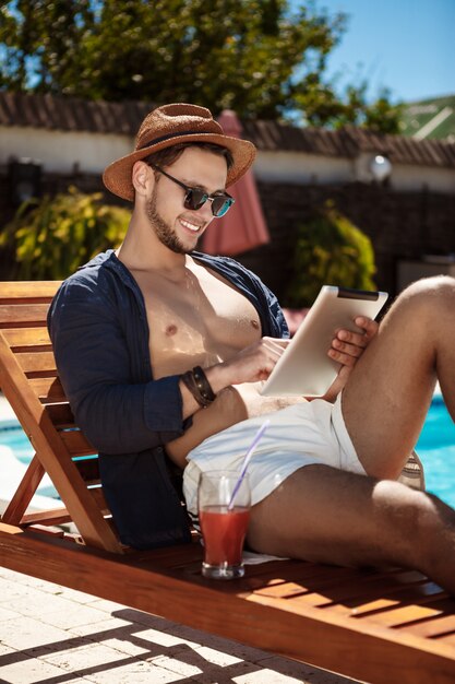 Junger schöner Mann, der Tablette betrachtet, die nahe Schwimmbad sitzt