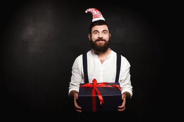 Junger schöner Mann, der Weihnachtsgeschenkbox über Schwarz hält.