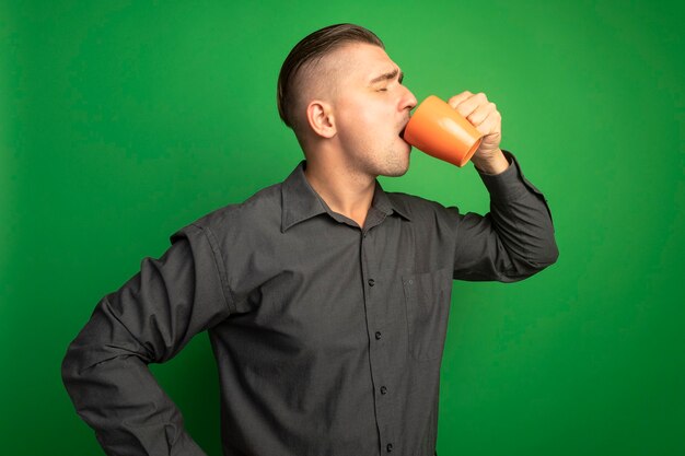 Junger schöner Mann im grauen Hemd, das Tee trinkt, der orange Becher hält