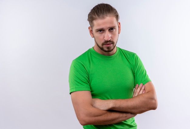 Junger schöner Mann im grünen T-Shirt, das mit verschränkten Armen steht und Kamera mit ernstem Gesicht über weißem Hintergrund betrachtet