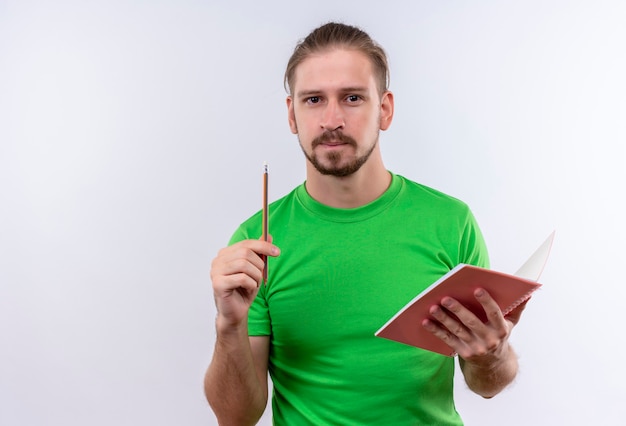 Junger schöner Mann im grünen T-Shirt, das Notizbuch hält, das mit Bleistift oben zeigt, eine große Idee, die über weißem Hintergrund steht