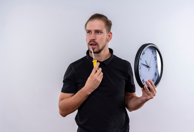 Junger schöner Mann im schwarzen Poloshirt, das Wanduhr und Schraubendreher hält, die mit nachdenklichem Ausdruck auf Gesicht stehen, das über weißem Hintergrund steht