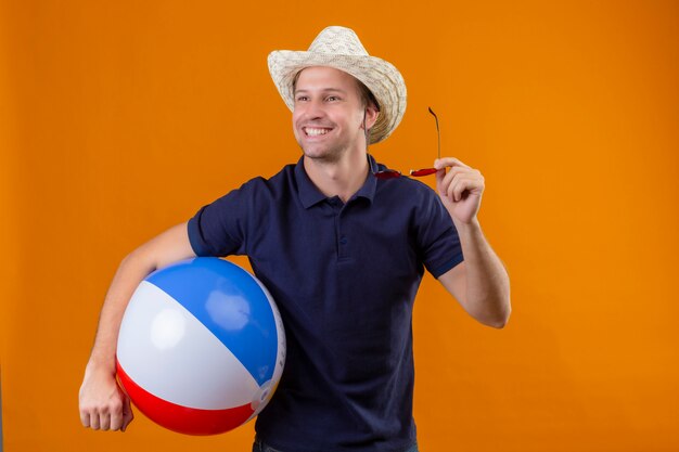 Junger schöner Mann im Sommerhut, der aufblasbaren Ball und die Sonnenbrille hält, die fröhlich mit glücklichem Gesicht stehen, das über orange Hintergrund steht